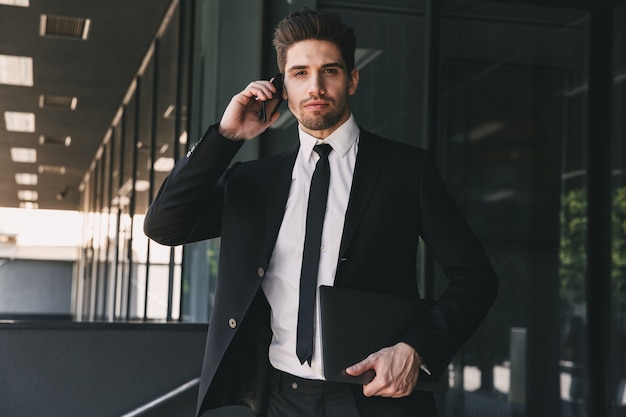 Portrait of european businessman dressed in formal suit standing outside glass building, and talking on mobile phone