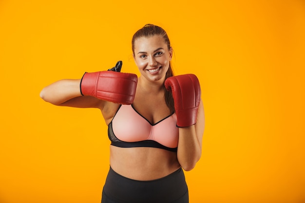 Ritratto di donna paffuta europea in reggiseno sportivo indossando guanti da boxe praticando, isolato su sfondo giallo