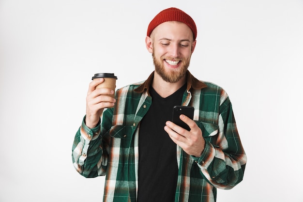 Portrait of european guy wearing plaid shirt holding paper cup with coffee and using mobile phone, while standing isolated over white background