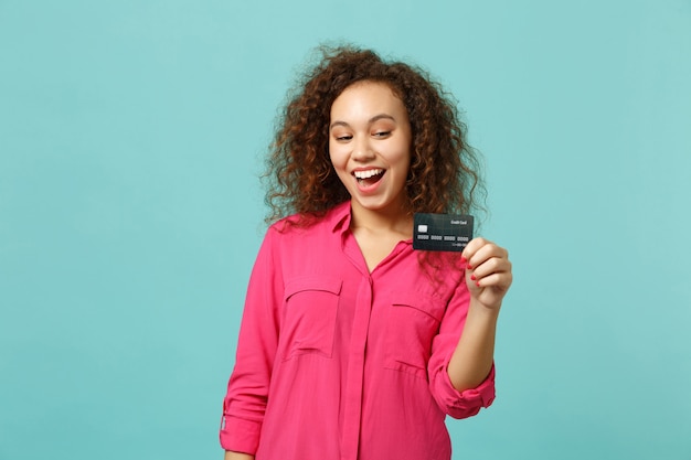 Photo portrait of excited african girl in pink casual clothes holding credit bank card isolated on blue turquoise wall background in studio. people sincere emotions, lifestyle concept. mock up copy space.