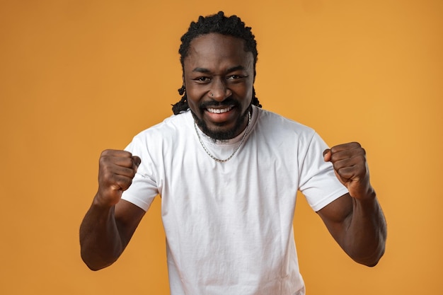 Portrait of excited african guy celebrating success with clenched fists against yellow background