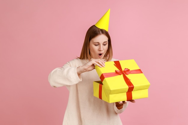 Portrait of excited amazed blond woman in party cone unpacking present box having pleasant surprise being shocked wearing white sweater Indoor studio shot isolated on pink background