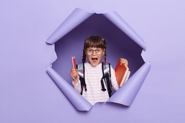 Portrait of excited amazed little girl in a striped shirt with textbooks in her hands and a backpack screaming happily rejoicing starting new school year