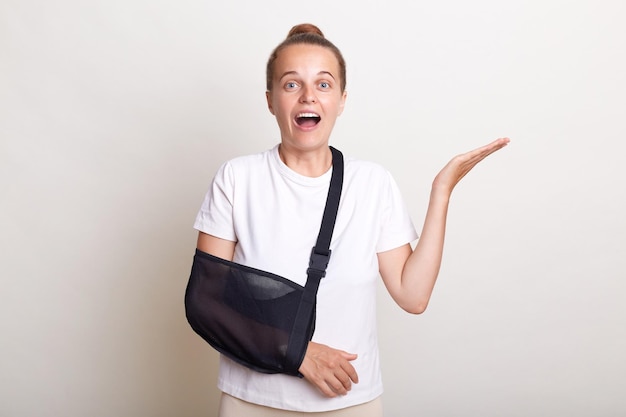 Photo portrait of excited amazed woman with bun hairstyle wearing casual t shirt posing isolated over white background female in arm sling after visiting surgeon being on sick leave