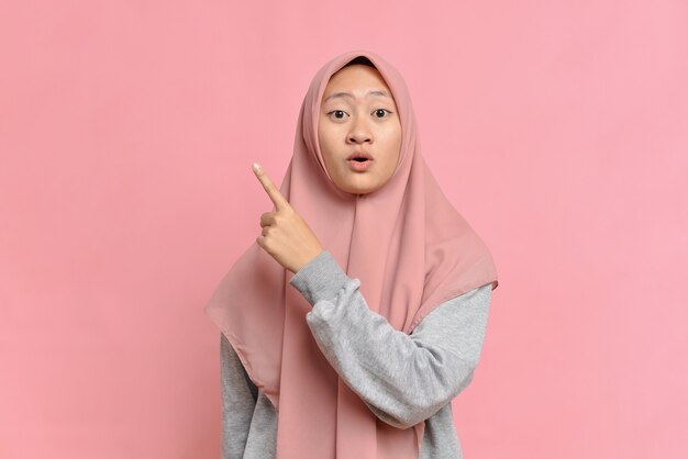 Portrait of Excited Asian beautiful young muslim woman pointing a copy space, isolated on pink background