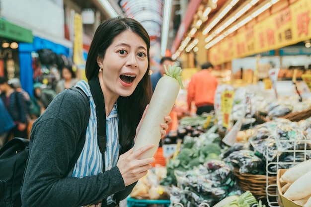Portrait of excited asian female tourist holding white radish\
and looking at camera with her mouth open at local stall selling\
fresh produce in kuromon ichiba market in osaka japan
