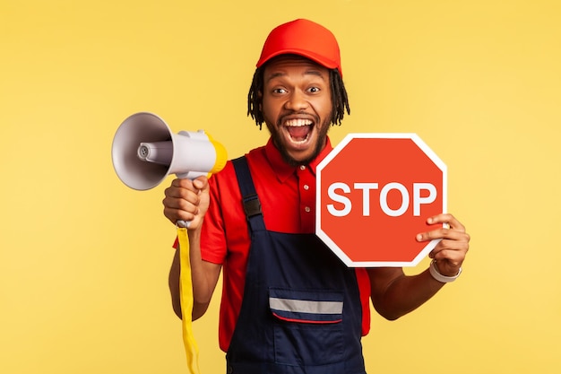 Portrait of excited bearded handyman wearing blue overalls and red Tshirt looking at camera holding red stop sign and megaphone in his hands Indoor studio shot isolated on yellow background