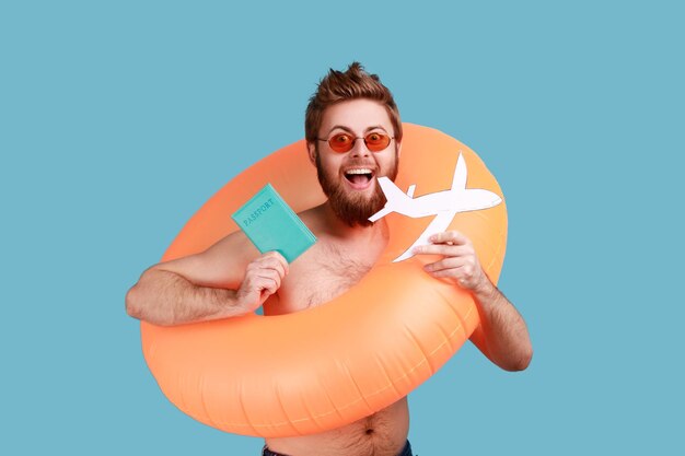 Portrait of excited bearded man standing with orange rubber ring, holding passport document, airplane mockup, expressing positive emotions. Indoor studio shot isolated on blue background.