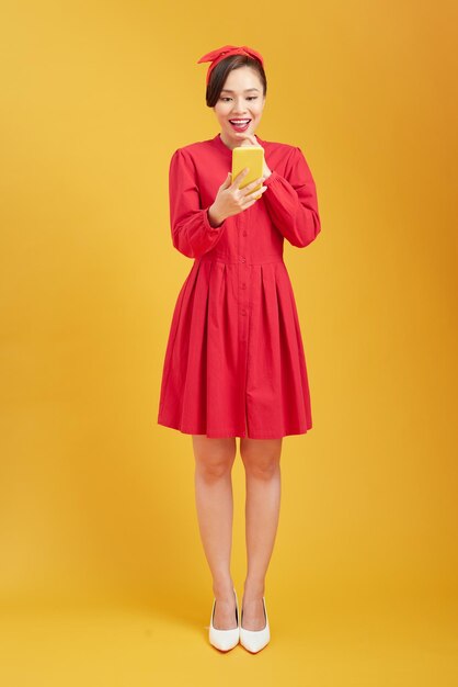 Portrait of an excited beautiful girl wearing dress holding mobile phone isolated over yellow background