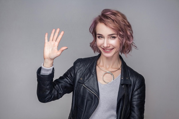 Portrait of excited beautiful girl with short hairstyle and makeup in black leather jacket standing, greeting and looking at camera with toothy smile. indoor studio shot, isolated on grey background.