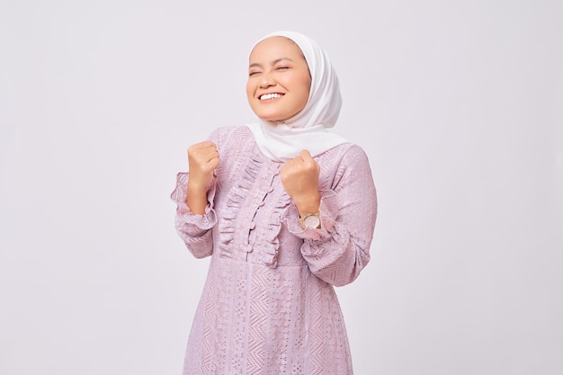 Portrait of excited beautiful young Asian Muslim woman wearing hijab and purple dress raising fists and celebrating victory on Ramadan isolated on white studio background