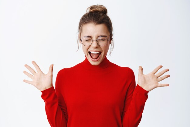 Portrait of excited blond woman in glasses, screaming from joy and happiness, raising hands up, celebrate victory, triumphing from success, white wall