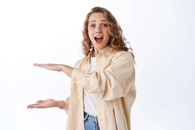 Portrait of excited blond woman showing big large size empty space logo on copy space standing amazed and looking at camera white background