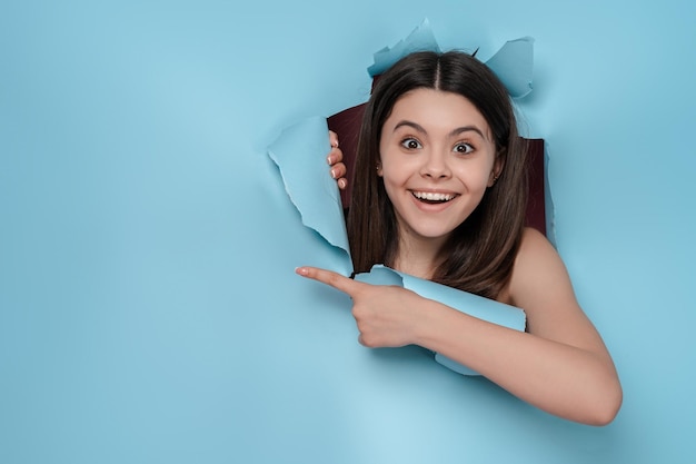 Photo portrait of excited brunette teenage girl looking over torn background pointing finger at copy space