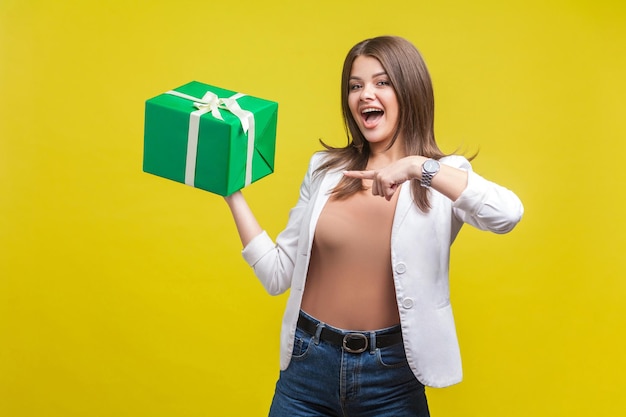 Portrait of excited brunette woman in jacket and jeans pointing at gift box in her hand looking at camera with open mouth and amazement pleasantly surprised indoor studio shot yellow background