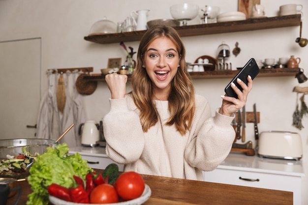 portrait of excited caucasian woman making winner gesture and using cellphone while cooking dinner in cozy kitchen