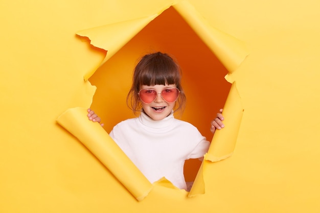 Ritratto di eccitata bambina affascinante con i capelli scuri che indossa un dolcevita bianco in posa in un buco strappato di muro di carta gialla in piedi con un'espressione facciale felice positiva