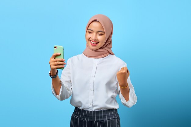 成功を祝う携帯電話で見ている興奮した陽気な若いアジアの女性の肖像画