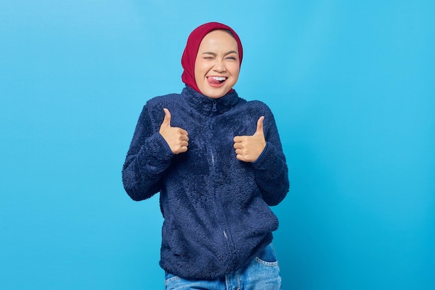 Портрет возбужденной жизнерадостной молодой азиатской женщины показывает палец вверх или знак одобрения на синем фоне