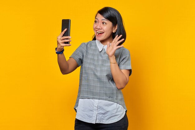 Portrait of excited cheerful young Asian woman using mobile phone take selfie over yellow background