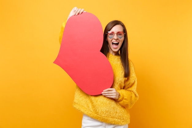 Portrait of excited cheerful young woman in fur sweater, heart glasses holding empty blank red heart isolated on bright yellow background. People sincere emotions, lifestyle concept. Advertising area.