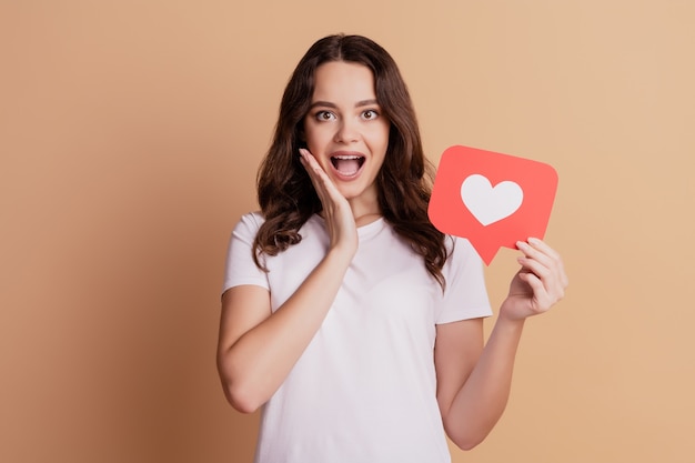 Portrait of excited crazy girl hand cheek open mouth hold notification heart icon isolated on beige background
