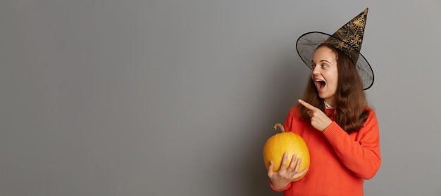 Photo portrait of excited crazy woman dressed in witch hat holding orange pumpkin and screaming isolated on gray background pointing away at mockup empty wall for commercial content place for adv
