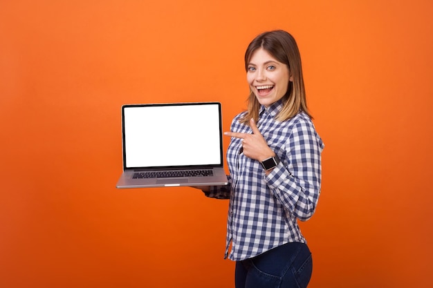 Portrait of excited cute woman with brown hair in checkered casual shirt standing pointing at laptop with blank screen and smiling place for ad indoor studio shot isolated on orange background