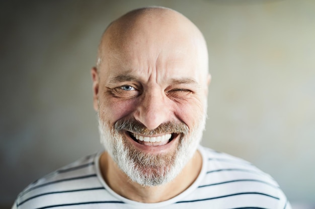 Portrait of excited emotional bald mature man with gray beard and mustache laughing cheerfully and winking at camera