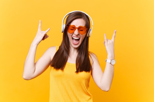 헤비메탈 록 기호를 보여주는 헤드폰으로 음악을 듣고 있는 주황색 안경을 쓴 흥분된 재미있는 소녀의 초상화, 노란색 배경에 격리된 복사 공간. 사람들이 진실한 감정 개념입니다. 광고 영역입니다.
