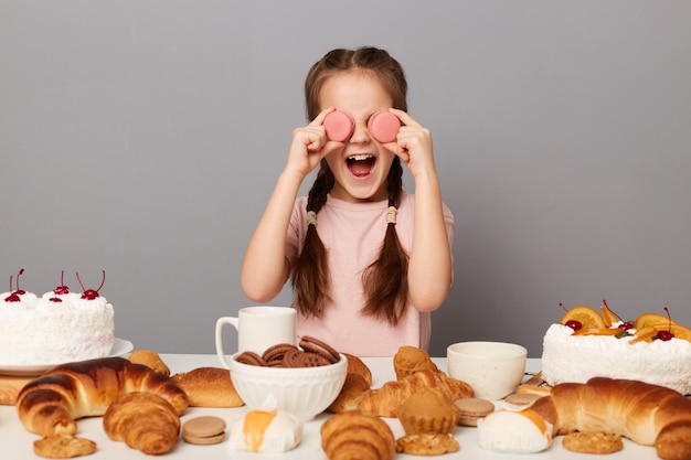 Портрет взволнованной забавной маленькой девочки с косичками, сидящей за столом на сером фоне, поедающей сладости, десерты, весело закрывающей глаза вкусными миндальными печеньями