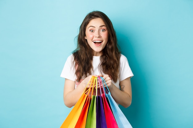 Portrait of excited girl shopper, holding shopping bags, buying in stores and smiling amazed, enjoy discounts, standing over blue background