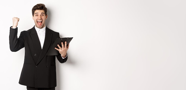 Portrait of excited handsome businessman in black suit holding clipboard and making fist pump achiev