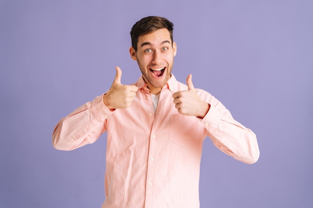 Портрет возбужденного красивого молодого человека, аплодирующего и указывающего пальцами, делающего двойной жест на розовом изолированном фоне в студии