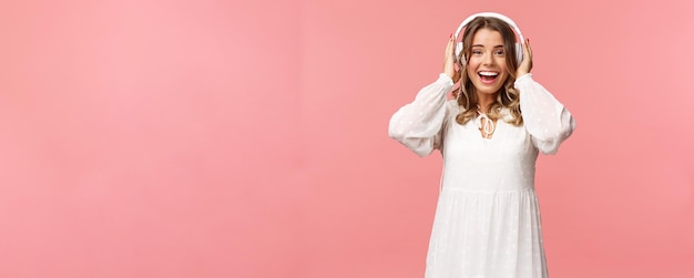 흰색 부드러운 드레스를 입고 헤드폰을 끼고 웃고 있는 흥분된 행복한 미녀의 초상화는 좋은 음질의 분홍색 배경에 매료된 카메라를 보고 놀라고 있습니다.
