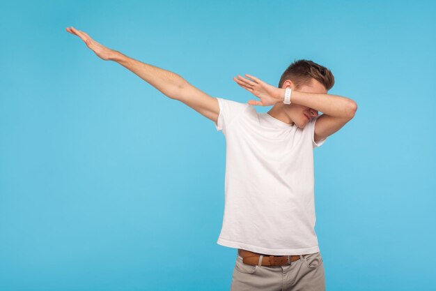 Portrait of excited happy man in white t-shirt showing dab dance, popular internet meme pose, celebrating success victory, dabbing trends concept. indoor studio shot isolated on blue background