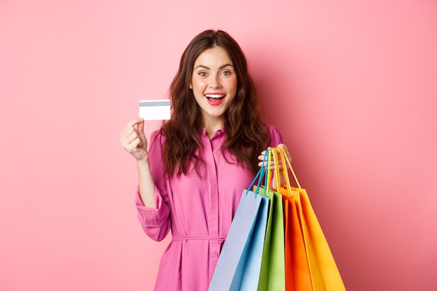 Foto ritratto di eccitato felice shopaholic, donna che tiene le borse della spesa e mostrando carta di credito in plastica, sorridendo stupito, in piedi contro il muro rosa.