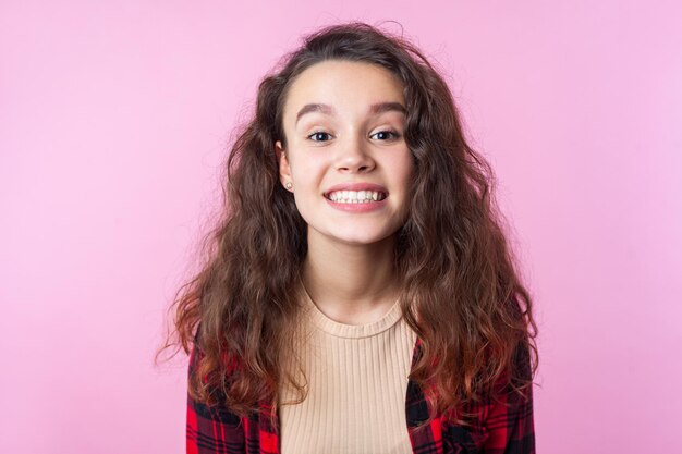 Portrait of excited happy teenage girl with curly brunette hair in casual shirt showing beautiful toothy smile at camera, white clean healthy teeth. indoor studio shot isolated on pink background