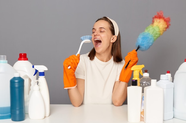 Portrait of excited happy woman wears headband and casual t\
shirt sitting among detergents holding cleaning brush as a\
microphone and singing having fun while tidy up isolated over gray\
background