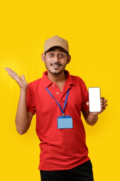 Photo portrait of a excited happy young delivery man in red t shirt and showing smartphone over yellow background.