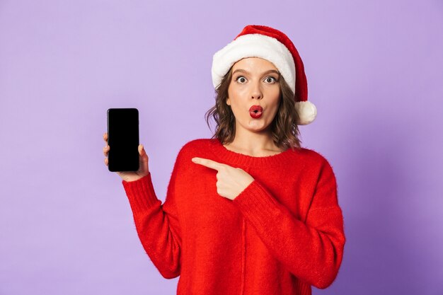 Ritratto di una giovane donna felice eccitata che porta il cappello di natale isolato sopra la parete viola che mostra la visualizzazione del telefono cellulare.