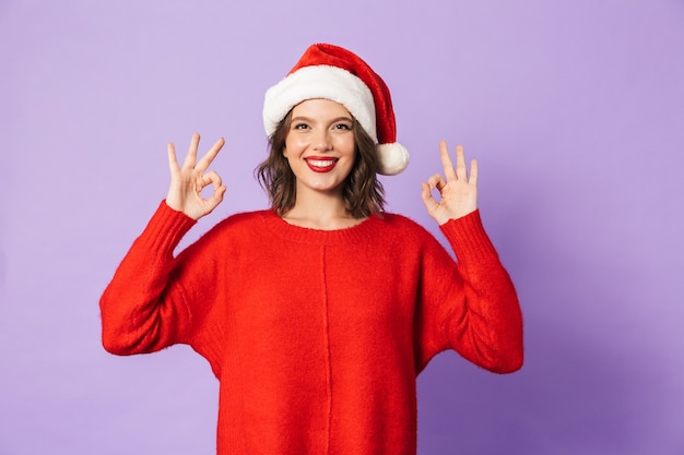 Ritratto di una giovane donna felice eccitata che porta il cappello di natale isolato sopra la parete viola che mostra il gesto giusto.