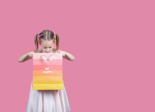 Портрет взволнованной маленькой девочки в платье и с красочными сумками для покупок, изолированными на розовом фоне с копировальным пространством.