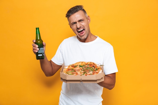 Ritratto di un uomo eccitato di 30 anni in maglietta bianca che beve birra e mangia pizza mentre si trova isolato sul giallo