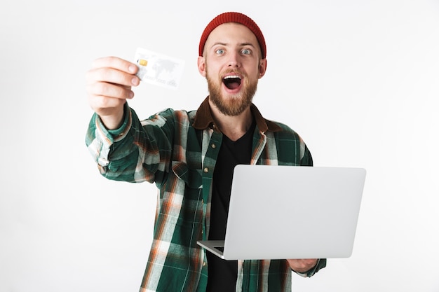 Portrait of excited man holding silver laptop and credit card, while standing isolated over white background