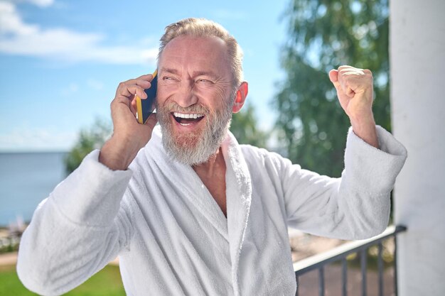 Portrait of an excited mature holidaymaker dressed in the white bathrobe talking on the smartphone