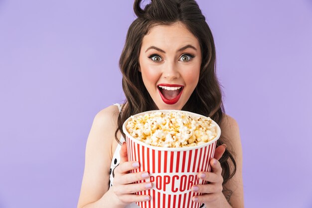 Portrait of excited pin-up woman in retro polka dot dress holding bucket with popcorn while watching movie isolated over violet wall