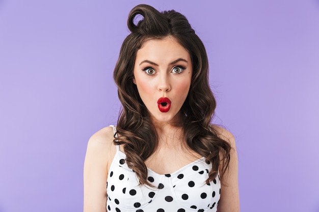 Portrait of excited pin-up woman in vintage polka dot dress smiling and rejoicing at camera isolated over violet wall