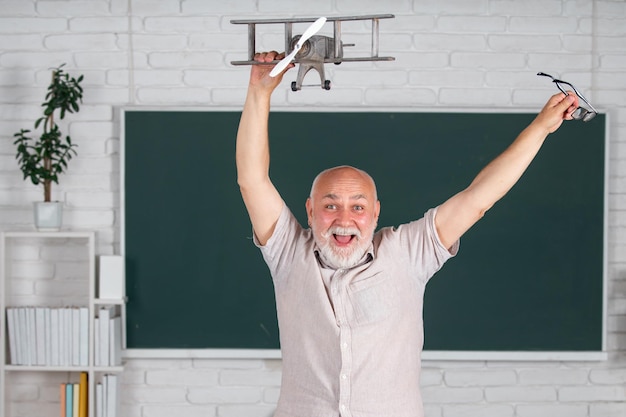 Portrait of excited senior teacher teaching line of high school students with computer laptop in classroom on blackboard Amazed aged male retired teacher with toy plane