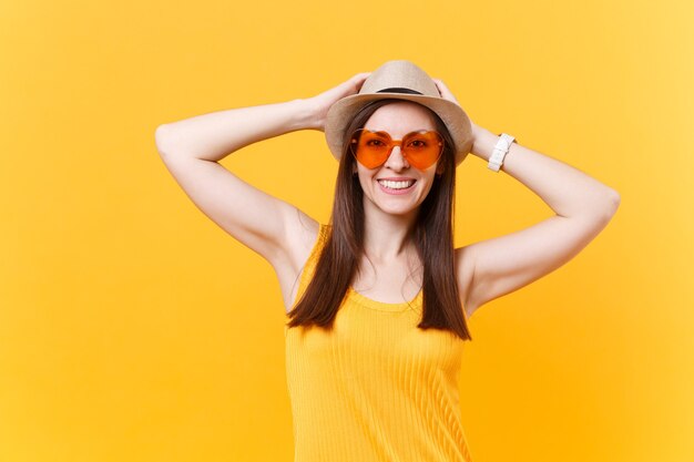 Ritratto di giovane donna sorridente eccitata in cappello estivo di paglia, occhiali arancioni mettono le mani sulla testa, copia spazio isolato su sfondo giallo. persone sincere emozioni, concetto di stile di vita. zona pubblicità.